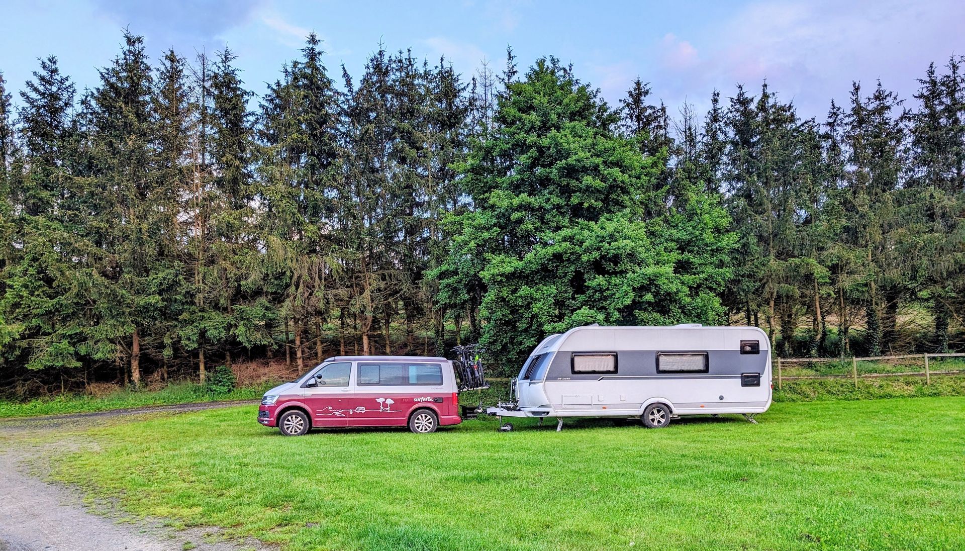 VW-Campingbus mit angehängtem Hobby-Wohnwagen auf einer Wiese vor dunklem Nadelwald in der Dämmerung.